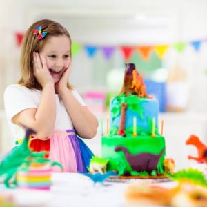 Kids birthday party. Dinosaur theme cake. Little girl blowing candles and opening gifts. Children event. Decoration for dinosaurs themed celebration.