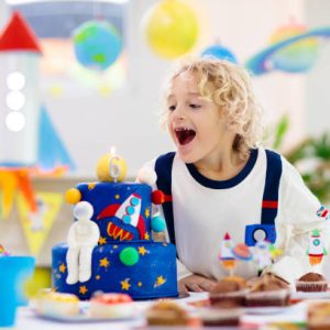 Kids space theme birthday party with cake and cupcakes. Rocket, solar system planet and spaceship decoration for child event. Little boy in astronaut costume blowing candles and opening presents.