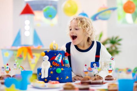 Kids space theme birthday party with cake and cupcakes. Rocket, solar system planet and spaceship decoration for child event. Little boy in astronaut costume blowing candles and opening presents.